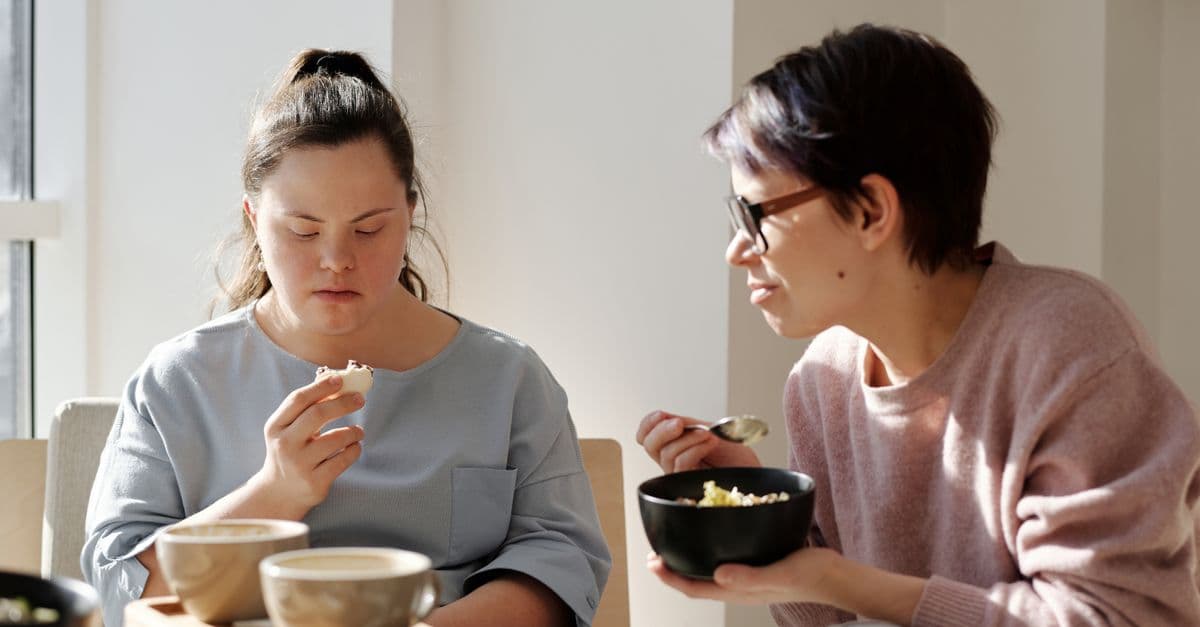 A young woman with Down syndrome sharing breakfast with her caregiver at a sunlit table.
